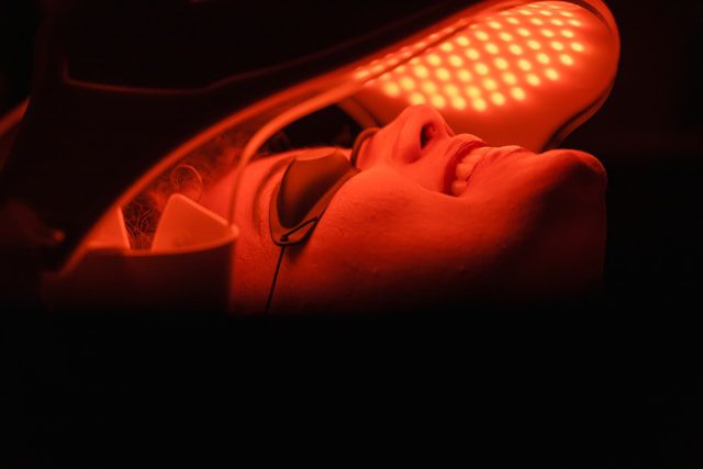 Person undergoing red light LED therapy wearing protective goggles, with face illuminated under a curved LED panel